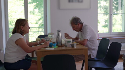 Obraz premium Senior couple eating breakfast together at home seated at dining table, enjoying morning meal with yogurt, coffee, and fruit near bright windows in cozy modern apartment