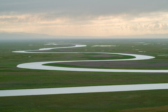 Aerial view of a winding river snaking through a vast green plain under a soft, hazy sky, Ba Yin Guo Leng Meng Gu Zi Zhi Zhou, Xin Jiang Wei Wu Er Zi Zhi Qu, China.