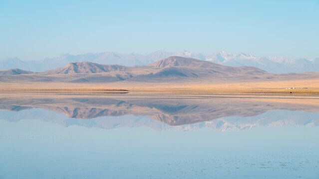 View of the serene lake mirroring the golden hills under a soft sky, with distant snowy peaks adding a touch of majesty, Karasaz, Almaty Region, Kazakhstan.