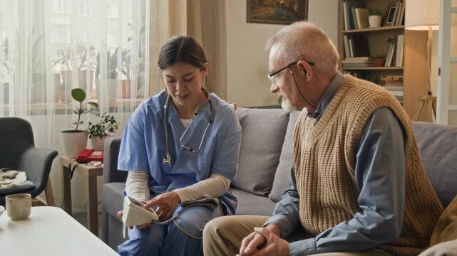 Young helpful nurse explaining how to use blood pressure monitor to elderly man, while sitting on couch in cozy retirement home