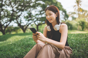 Happy asian woman wearing headphones, sitting on green grass, listening to music and watching content on her mobile phone, A digital lifestyle, relaxation in nature