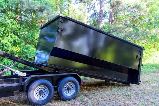 Black dumpster is placed on trailer in green outdoor location with trees around