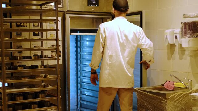Baker placing trays with raw dough buns into a professional refrigeration cabinet, controlling fermentation and preparation process in an industrial bakery kitchen
