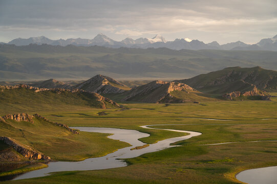 Aerial view of a winding river snaking through a lush valley, framed by rugged hills and distant snow-capped mountains, Ba Yin Guo Leng Meng Gu Zi Zhi Zhou, Xin Jiang Wei Wu Er Zi Zhi Qu, China.