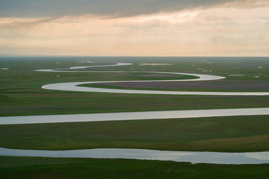 Aerial view of the winding river snaking through the expansive green meadows, bathed in the soft glow of the setting sun, Ba Yin Guo Leng Meng Gu Zi Zhi Zhou, Xin Jiang Wei Wu Er Zi Zhi Qu, China.