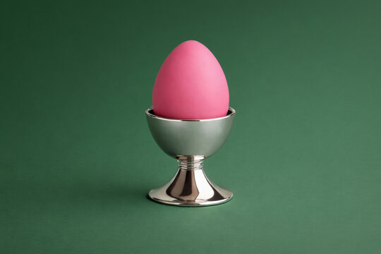 Pink egg placed in metallic egg cup against deep green background. Minimal still life with strong color contrast, modern seasonal styling. Easter concept.