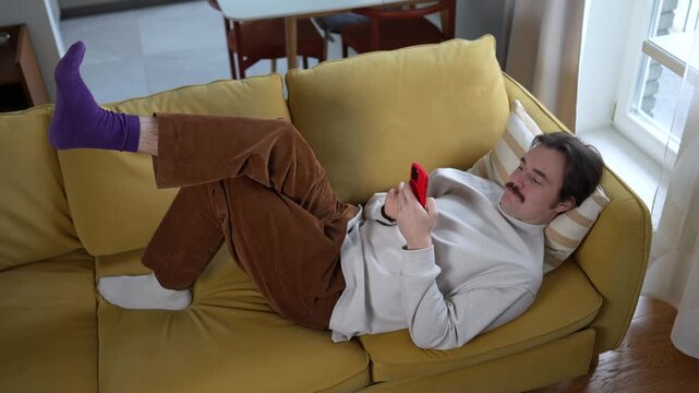 Disinterested lazy man with phone lying on couch surfing online web, procrastination at home. Unmotivated male with social media addiction on smartphone scrolling internet, look at screen virtual life