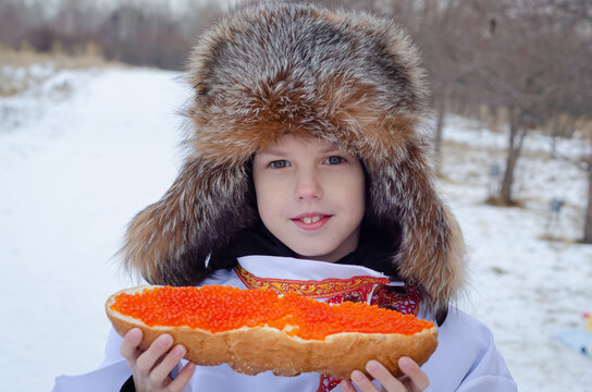 Portrait of a boy in a fur hat and a Russian shirt: a large sandwich with red caviar is a symbol of the winter feast and traditional cuisine of Russia.