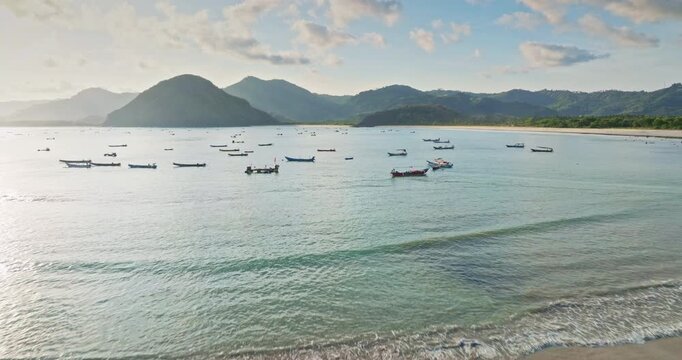 Selong Belanak Beach, Lombok: traditional fishing boats in calm bay with gentle waves and sandy shore, clear blue water and green hills under a sunny sky. Tropical nature landscape. Aerial view flight