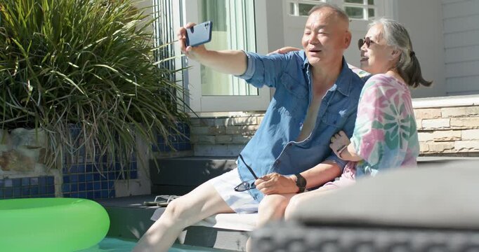Mature Asian couple posing at pool edge while man holding smartphone and sunglasses, capturing kiss