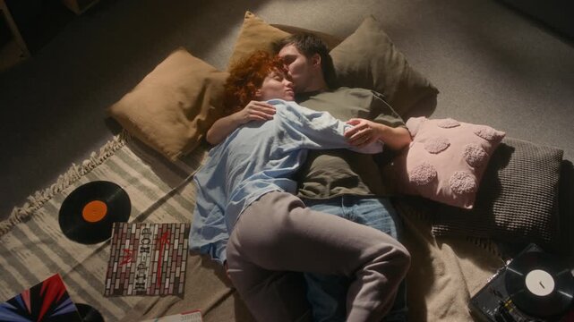 Cinematic top view shot of happy young couple lying on floor over pile of pillows and cuddling in soft sunlight listening to music with retro record player and records scattered around, copy space