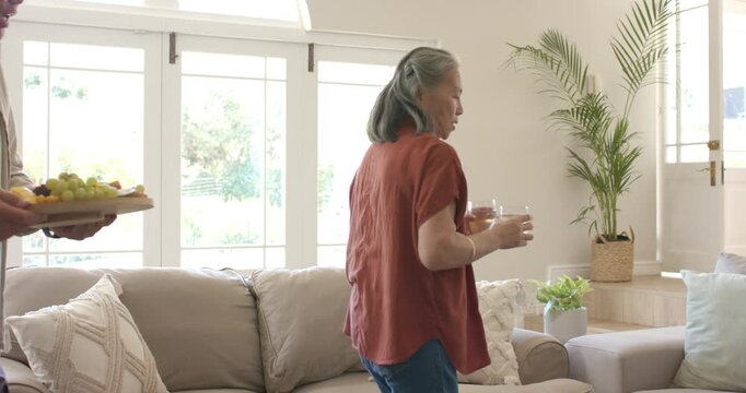 Asian senior couple bringing refreshments walking along sofa balancing glasses placing fruit tray
