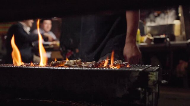 Slow motion of a cook grilling skewers over open fire at a street yatai in Fukuoka, Japan. Flames rise as people wait in the background during a busy evening.