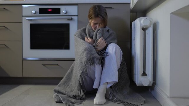 Frozen, distressed young woman sitting on floor wrapped in blanket near heater radiator shivering from cold. Lack of heating at home. Central heating failure, winter coldness indoor, utility crisis.