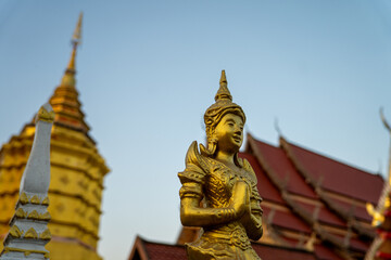 Fototapeta premium Golden angel statue in praying gesture with temple pagoda at sunset sky, sacred Thai sculpture representing faith, devotion, Wat Luang, Chiang Khong, Chiang Rai, Thailand