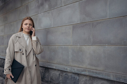 Young adult business woman has a talk by mobile phone going on city street in front of stone wall