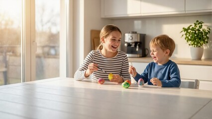Fototapeta premium Two laughing children painting colorful Easter eggs at kitchen table. Happy siblings enjoying holiday traditions in sunlit home. Creative spring activity. Copy space for text
