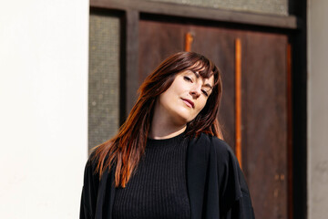 Fototapeta premium Confident woman with long brown hair wearing a black sweater poses against a rustic wooden door, showcasing a blend of modern style and natural beauty in an urban setting