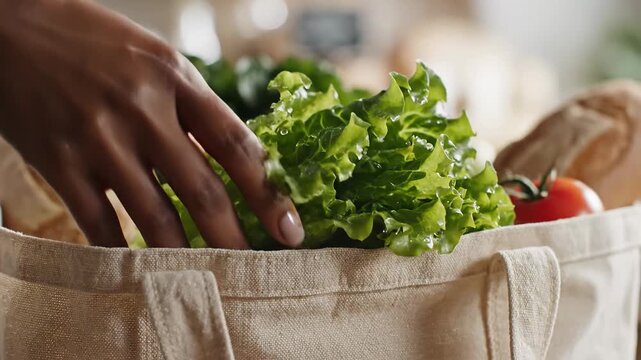 Hands placing fresh wet lettuce into a reusable grocery bag with bread and tomatoes