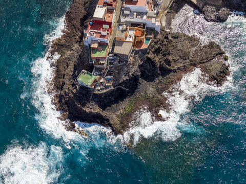 Aerial view of the rugged coastline with fisher village whitewashed buildings meet the turquoise ocean, against the dark volcanic rock, Caleta de Arriba, Gran Canaria, Spain.