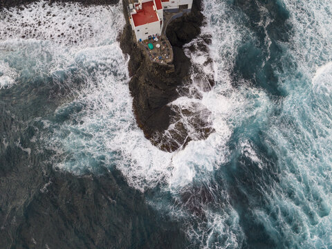Aerial view of white buildings clinging to a rugged, dark cliff, as turquoise waves crash around its base, a dance of color and form, Gran Canaria, Moya, Canary Islands, Spain.