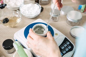 Hands stir gray cosmetic paste in small bowl on digital scale surrounded by laboratory glassware and jars. Small-batch manufacturing, slow beauty movement, artisan production.