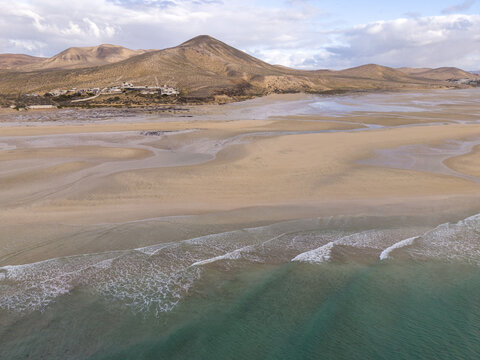 Aerial view of the vast sandy beach meeting the turquoise ocean under a cloudy sky, with mountains in the backdrop, Jand&Atilde;&shy;a Natural Park, Fuerteventura, Canary Islands, Spain.