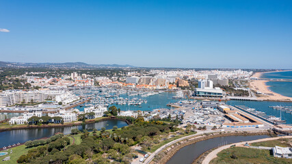 Obraz premium A wide view shows Vilamoura marina with boats docked, people enjoying the area, and buildings lining the waterfront under a blue sky Aerial