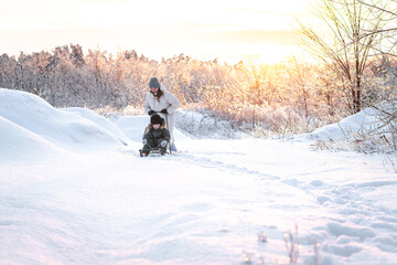 A young mother takes her son sledding in a snowy area against the backdrop of a sunset - a concept of winter, motherhood, and winter activities.