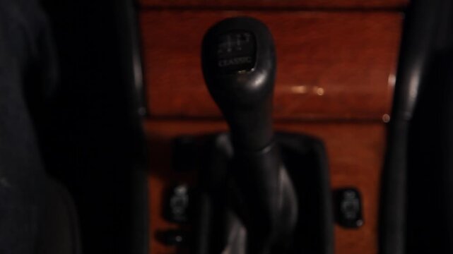Close up of driver's hand shifting manual gear stick in car. The black knob features the word classic and the interior has elegant wood trim panels.

