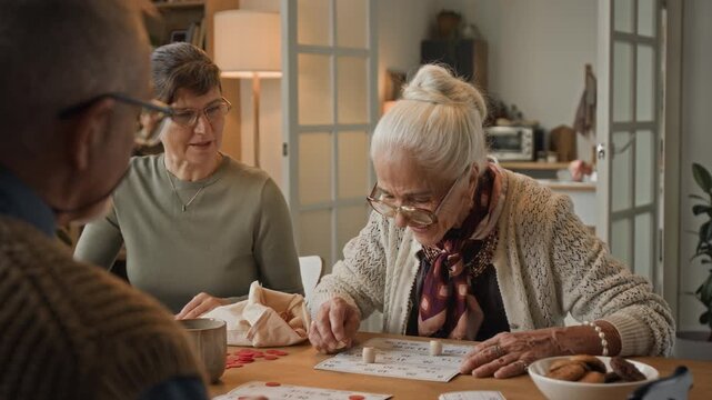 Over the shoulder shot of three cheerful seniors playing bingo game and communicating, sitting at table in retirement home