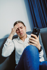 Woman sitting on couch holding phone with hand on forehead while looking stressed in indoor setting