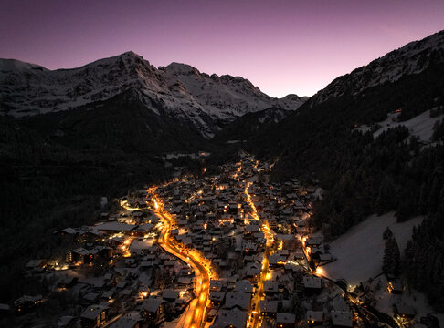 Aerial view of a village nestled between dark forests and snow-capped mountains under a dusky sky, glowing with warm lights, Champ&Atilde;&copy;ry, Valais, Switzerland.