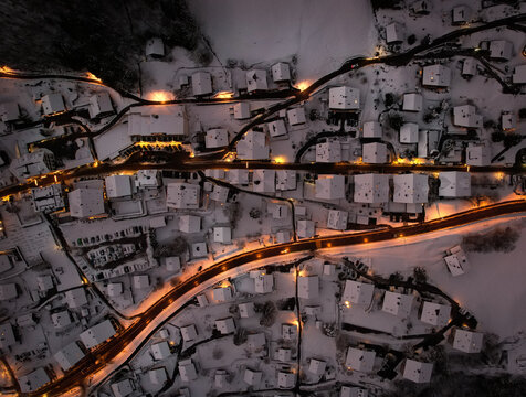 Aerial view of a snow-blanketed village with golden lights tracing roads and illuminating rooftops, creating a warm glow against the cold winter landscape, Champ&Atilde;&copy;ry, Valais, Switzerland.