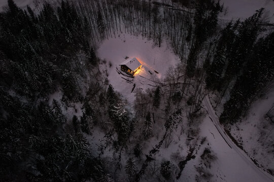Aerial view of a warmly lit cabin nestled in the snow-covered forest, a beacon of light against the dark trees, Champ&Atilde;&copy;ry, Valais, Switzerland.