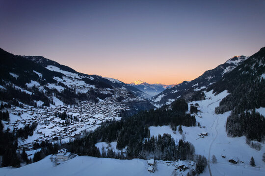 Aerial view of a valley blanketed in snow with scattered chalets nestled among the trees, framed by majestic mountains under a pastel sky, Champ&Atilde;&copy;ry, Valais, Switzerland.