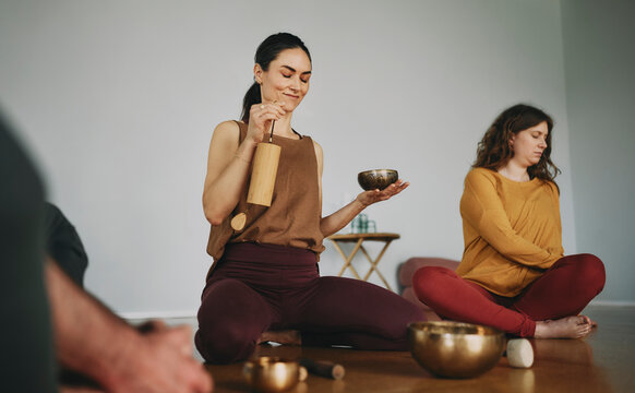 Female yoga instructor beginning a meditation class using a singing bowl and chime with a group of students sitting on the floor of her studio