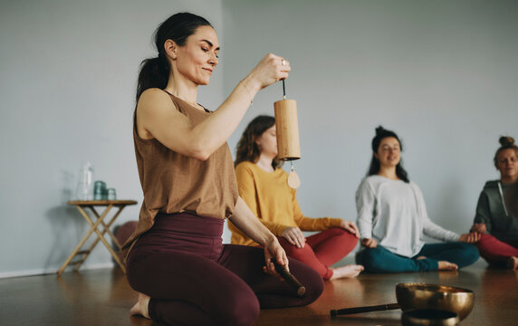 Female yoga teaching beginning a meditation class using a chime and singing bowl with a group of students sitting on the floor of her studio