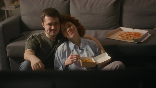 Front view shot of happy young couple watching TV together and enjoying fast food while sitting on floor in cozy home setting, copy space