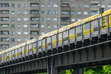 Yellow Berlin U-Bahn Train on Elevated Steel Bridge with Brutalist Plattenbau Apartment Building Background Urban Transport Architecture Scene