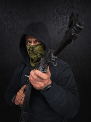 Naklejka premium Armed man in black hoodie and camouflage mask holding a rifle against a dark background, representing security, tactical concept, or soldier.