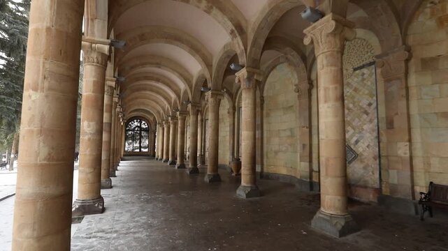 Walking inside an ancient stone arcade gallery with columns and arches. Winter view with snow outside large windows. Detailed mosaic walls and decorative stone vases line the corridor.

