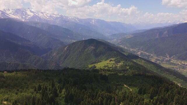 Aerial drone view glides forward and downward over conifer forests, alpine meadows, farmhouses, and switchback roads in Svaneti, Georgia, toward snow capped peaks.