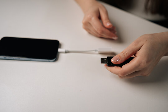Closeup view of unrecognizable woman connecting usb flash drive to smartphone through adapter cable, facilitating data transfer and expanding mobile storage sitting on clean white table.