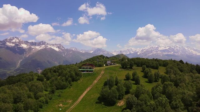 Aerial drone shot tracks a grassy ridge toward lift poles and buildings in Svaneti, Georgia, with Ushba like peaks, deep forests, alpine meadows, and midday cumulus clouds.