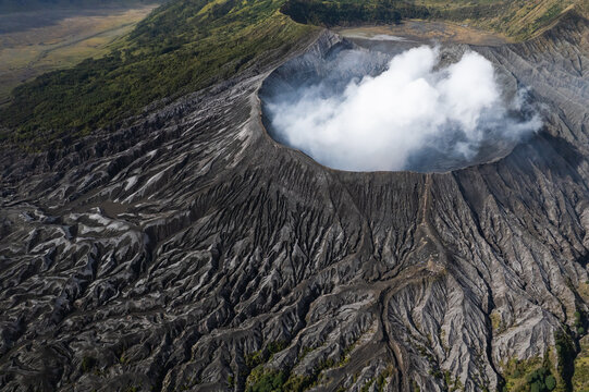 Aerial view of a brooding volcano, its dark, textured slopes contrasting sharply with the ethereal white plume rising from its caldera, Bromo, East Java, Indonesia.