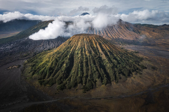 Aerial view of mist-shrouded Mount Bromo, with its smoky crater and textured slopes, contrasts against the dark volcanic earth, creating a scene of dramatic beauty, Probolinggo, East Java, Indonesia.