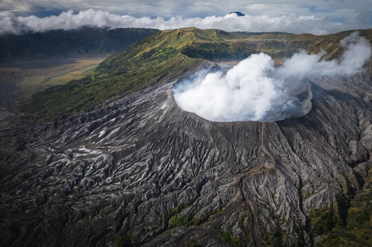 Aerial view of the volcanic crater of Mount Bromo steaming under a cloudy sky, its slopes textured in stark contrast to the green hills, Probolinggo, East Java, Indonesia.