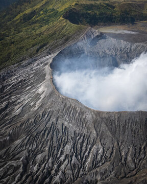 Aerial view of the dramatic, textured landscape of Mount Bromo with wisps of white smoke rising from the crater against the backdrop of green vegetation, Probolinggo, East Java, Indonesia.