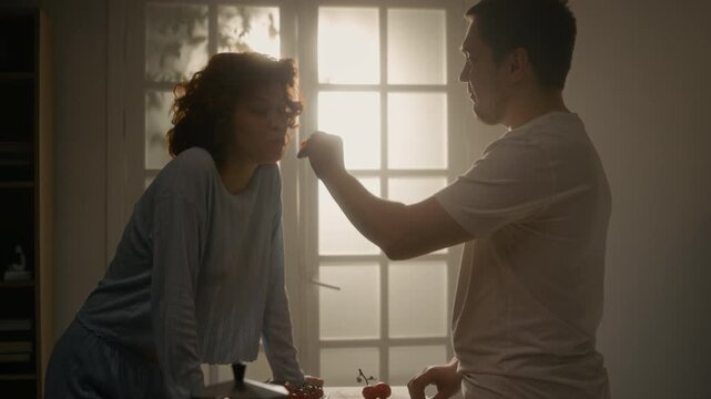Cinematic side view shot of happy young couple cooking together in morning sharing bites of food, scene backlit by soft sunlight